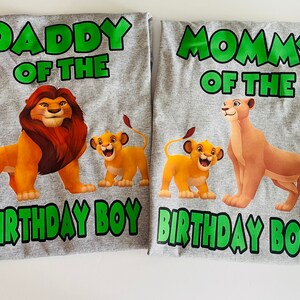 May include: Two gray t-shirts with cartoon lion family graphics. One shirt says "Daddy of the Birthday Boy" with a lion and cub. The other says "Mommy of the Birthday Boy" with a lioness and cub. Green text.