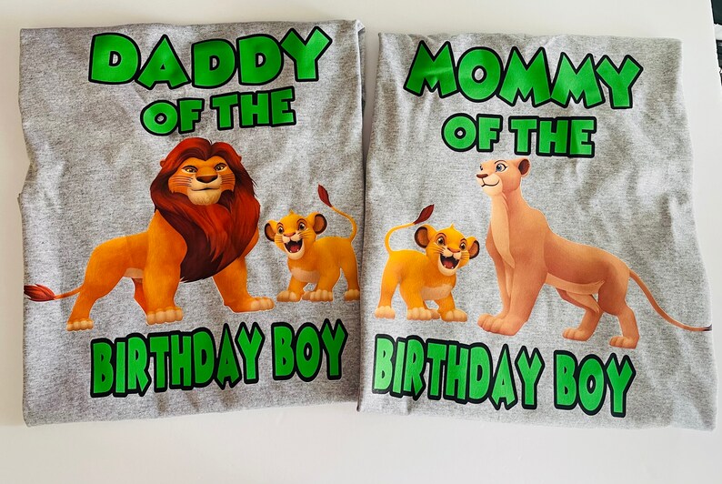 May include: Two gray t-shirts with cartoon lion family graphics. One shirt says "Daddy of the Birthday Boy" with a lion and cub. The other says "Mommy of the Birthday Boy" with a lioness and cub. Green text.