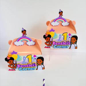 May include: Two peach-colored favor boxes with a rainbow, clouds, and stars design. Each box features a black girl with a pink dress and a birthday hat holding balloons. The boxes have the text "Kenzie's Corner" and the number "1" on them.
