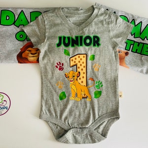 May include: A gray baby bodysuit with a Simba graphic, the number 1, and the word "JUNIOR" in green. The bodysuit is part of a set with a matching shirt that says "DADDY OF THE". Paw prints and leaves decorate the bodysuit.
