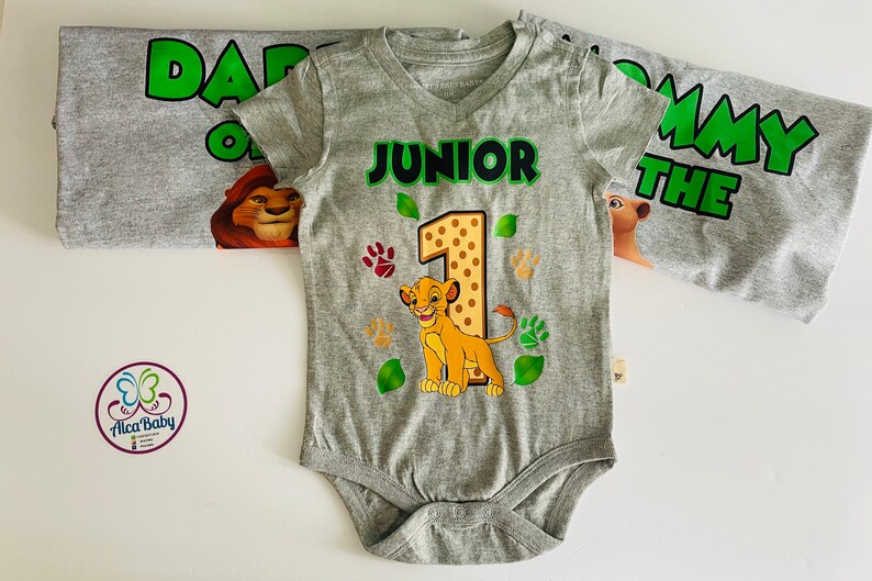 May include: A gray baby bodysuit with a Simba graphic, the number 1, and the word "JUNIOR" in green. The bodysuit is part of a set with a matching shirt that says "DADDY OF THE". Paw prints and leaves decorate the bodysuit.