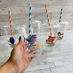 May include: Four clear plastic cups with colorful straws and Finding Nemo themed stickers. The stickers feature characters from the movie, including Dory, Nemo, and a shark. The cups are labeled "Finding Luka 3".