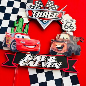 May include: A red and black cake topper with a checkered flag design, featuring the characters Lightning McQueen and Mater from the Disney Pixar Cars movie. The topper also includes a banner with the text "Kai & Calvin" and a sign that says "Three" and "Route 66".
