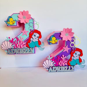May include: Two pink and purple numbers, 2 and 3, decorated with a mermaid, a starfish, seashells, and coral. The numbers are topped with a pink flower and have the name "Adrielly" written in silver glitter.