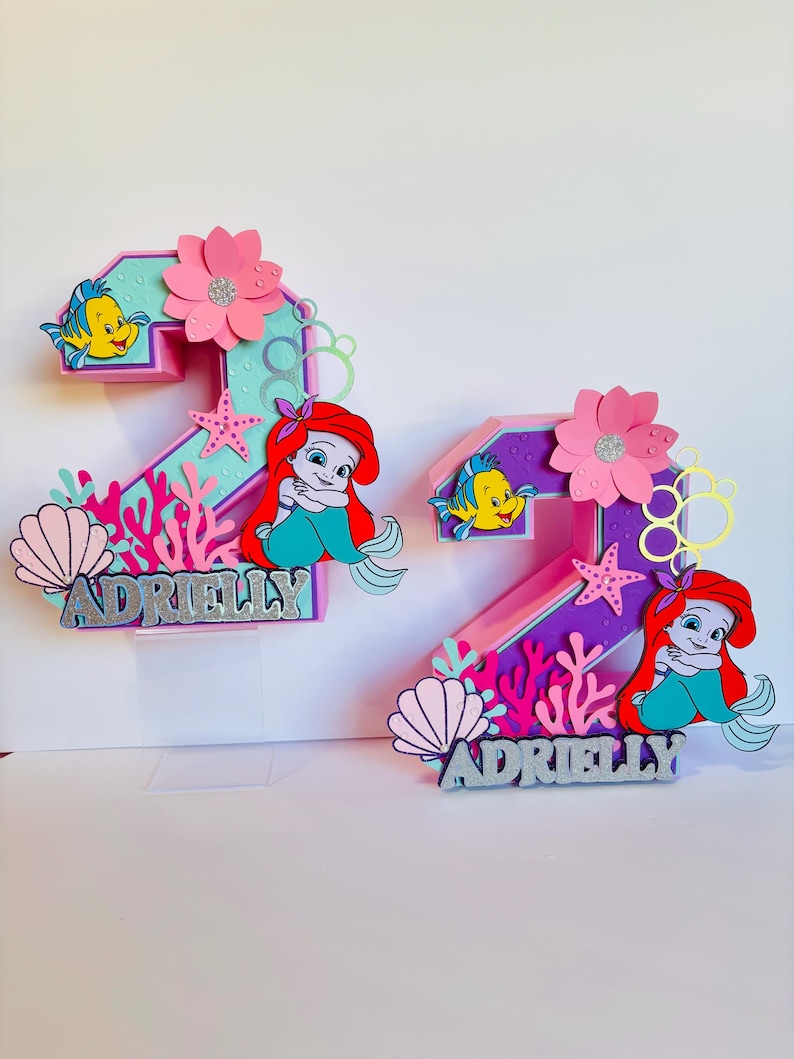 May include: Two pink and purple numbers, 2 and 3, decorated with a mermaid, a starfish, seashells, and coral. The numbers are topped with a pink flower and have the name "Adrielly" written in silver glitter.