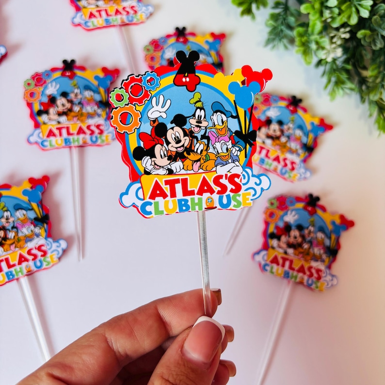 Peut inclure: D&eacute;corations de g&acirc;teau color&eacute;es avec Mickey Mouse, Minnie Mouse, Donald Duck, Dingo et Pluto. Les d&eacute;corations portent l'inscription "ATLAS'S CLUBHOUSE" et sont fix&eacute;es sur des b&acirc;tonnets en plastique transparents. Le design comprend des engrenages, des ballons et un fond de ciel bleu.