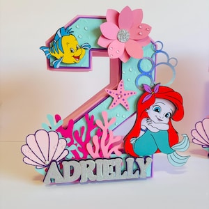 May include: A pink and blue number two cake topper with a mermaid, a starfish, coral, seashells, and the name "Adrielly" in silver glitter.
