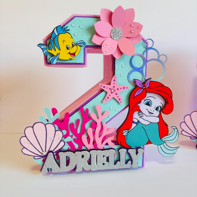 May include: A pink and blue number two cake topper with a mermaid, a starfish, coral, seashells, and the name "Adrielly" in silver glitter.