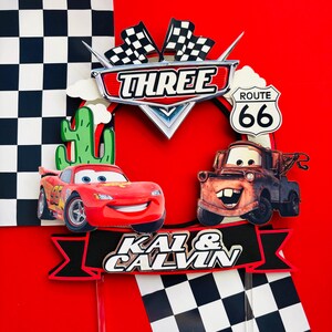 May include: A red and black cake topper featuring Lightning McQueen and Mater from the Cars movie. The topper includes a checkered flag, a cactus, and the text "THREE", "ROUTE 66", and "KAI & CALVIN".