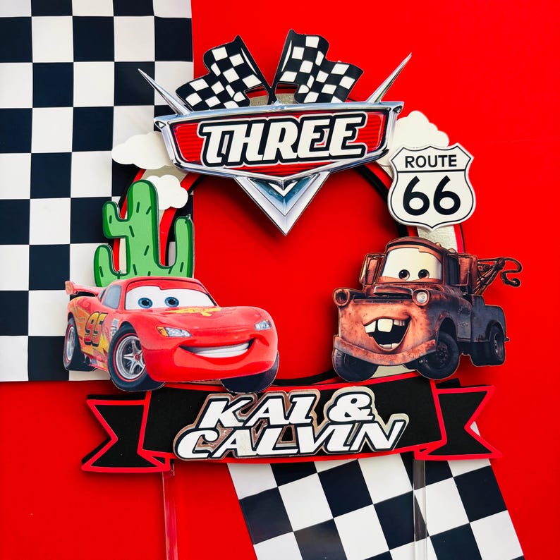 May include: A red and black cake topper featuring Lightning McQueen and Mater from the Cars movie. The topper includes a checkered flag, a cactus, and the text "THREE", "ROUTE 66", and "KAI & CALVIN".