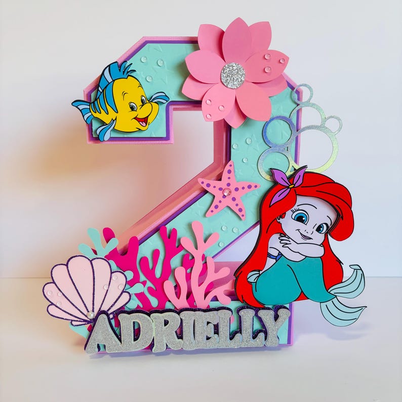 May include: A pink and purple number two cake topper featuring a mermaid, a seashell, coral, and a starfish. The topper also includes a yellow fish with a purple tail and the name "Adrielly" in silver glitter letters.
