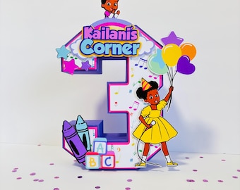 May include: A colorful number three cake topper with the text "Kailani's Corner". The topper features a cartoon girl holding balloons, musical notes, crayons, and ABC blocks. The design includes purple, yellow, blue, and pink accents.