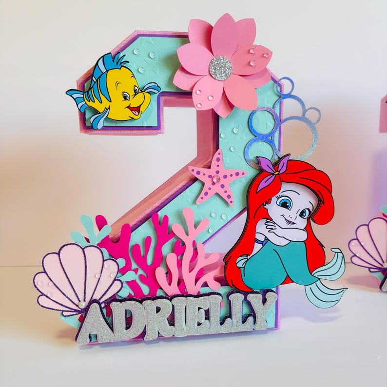 May include: A pink and blue 3D number two with a mermaid, seashells, coral, and a starfish. The number is decorated with glitter and the name "Adrielly" is written in silver glitter.