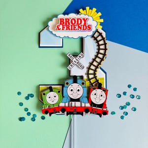 May include: A colorful birthday cake topper featuring the number 2, three cartoon train engines, and the text "BRODY & FRIENDS." The design includes a railroad crossing sign, a cloud, and a sun. The topper is decorated with blue and green sequins.