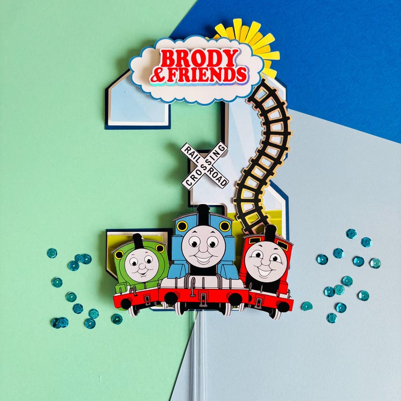 May include: A colorful birthday cake topper featuring the number 2, three cartoon train engines, and the text "BRODY & FRIENDS." The design includes a railroad crossing sign, a cloud, and a sun. The topper is decorated with blue and green sequins.