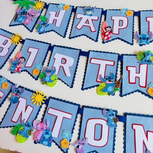 Puede incluir: Una pancarta azul con las palabras "HAPPY BIRTHDAY" en letras blancas. Decorada con im&aacute;genes de Stitch y flores tropicales. Ideal para una fiesta de cumplea&ntilde;os.