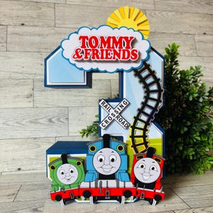 May include: A colorful, layered number 2 cake topper featuring Thomas the Tank Engine and friends. The design includes a blue number 2, a cloud with the text "TOMMY & FRIENDS", a railroad crossing sign, a sun, and a train track.