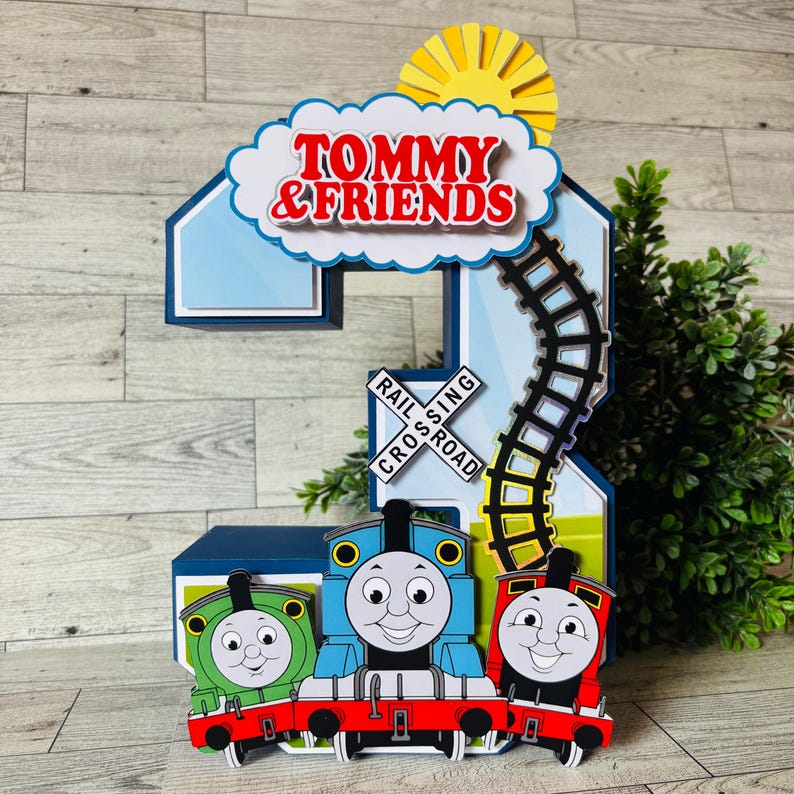 May include: A colorful, layered number 2 cake topper featuring Thomas the Tank Engine and friends. The design includes a blue number 2, a cloud with the text "TOMMY & FRIENDS", a railroad crossing sign, a sun, and a train track.
