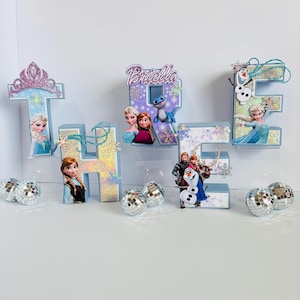 Pode incluir: Uma exibi&ccedil;&atilde;o decorativa com as letras "THE" e o nome "Briella", em azul claro e prata. As letras s&atilde;o adornadas com personagens do filme Frozen, flocos de neve e bolas de discoteca, criando um ambiente festivo.
