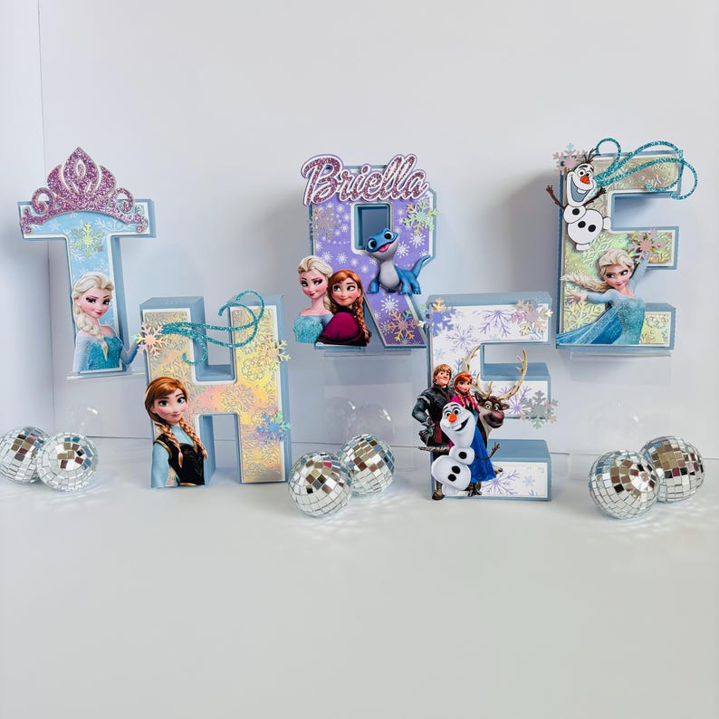 Pode incluir: Uma exibi&ccedil;&atilde;o decorativa com as letras "THE" e o nome "Briella", em azul claro e prata. As letras s&atilde;o adornadas com personagens do filme Frozen, flocos de neve e bolas de discoteca, criando um ambiente festivo.