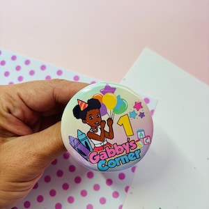 Pode incluir: Um botão redondo com uma ilustração de desenho animado de uma menina segurando balões, usando um chapéu de festa e o número 1. O botão tem o texto "Gabby's Corner" e imagens de lápis de cor e estrelas. O botão é segurado em uma mão.