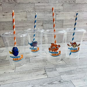 May include: Four clear plastic cups with orange and white striped straws. Each cup has a Finding Nemo themed sticker with a different character: Dory, a shark, Marlin, and a sea turtle. The stickers all say "Finding Luka 3".