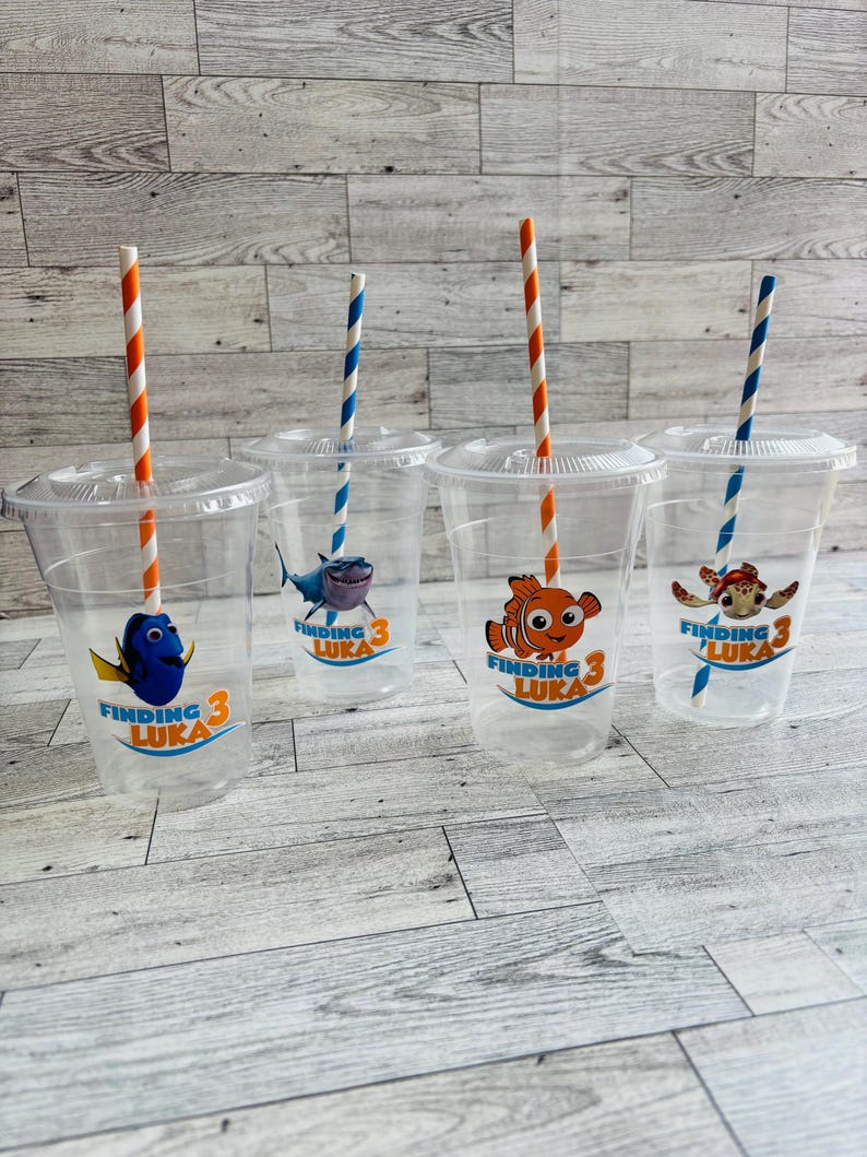May include: Four clear plastic cups with orange and white striped straws. Each cup has a Finding Nemo themed sticker with a different character: Dory, a shark, Marlin, and a sea turtle. The stickers all say "Finding Luka 3".