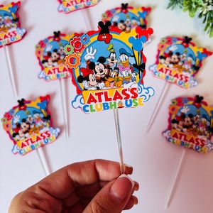 Peut inclure: D&eacute;corations de cupcakes color&eacute;es avec Mickey Mouse, Minnie Mouse, Dingo, Donald Duck et Pluto. Les d&eacute;corations ont un fond bleu avec le texte "ATLAS'S CLUBHOUSE". Chaque d&eacute;coration est fix&eacute;e &agrave; un b&acirc;tonnet en plastique transparent.