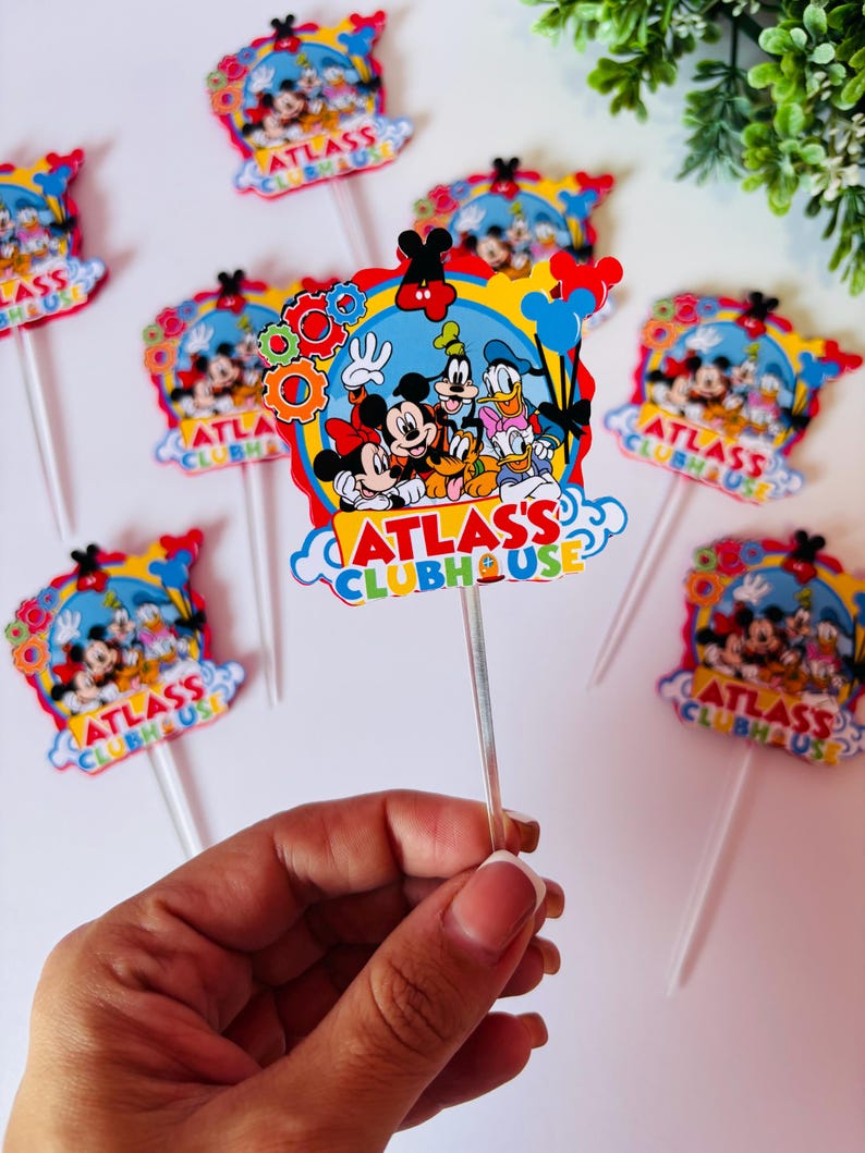 Peut inclure: D&eacute;corations de cupcakes color&eacute;es avec Mickey Mouse, Minnie Mouse, Dingo, Donald Duck et Pluto. Les d&eacute;corations ont un fond bleu avec le texte "ATLAS'S CLUBHOUSE". Chaque d&eacute;coration est fix&eacute;e &agrave; un b&acirc;tonnet en plastique transparent.