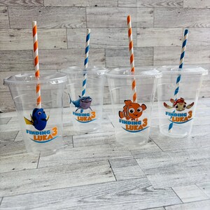 May include: Four clear plastic cups with Finding Nemo themed designs. Each cup has a different character from the movie, including Dory, Marlin, and Crush. The cups have orange and blue striped straws.