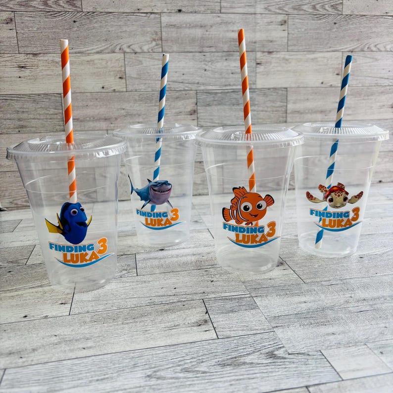 May include: Four clear plastic cups with Finding Nemo themed designs. Each cup has a different character from the movie, including Dory, Marlin, and Crush. The cups have orange and blue striped straws.