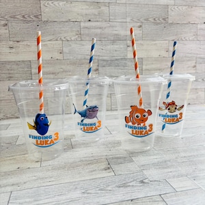 May include: Four clear plastic cups with Finding Nemo themed designs and orange and white striped straws. The designs feature Dory, Marlin, a shark, and a turtle. Each cup has the text "Finding Luka 3" printed on it.