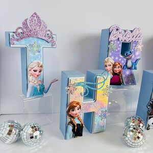 Pode incluir: Letras de cart&atilde;o azul claro que soletram "T", "4" e "H", decoradas com imagens de personagens do filme Frozen, flocos de neve e glitter. A letra "T" &eacute; encimada por uma tiara brilhante. Bolas de discoteca prateadas est&atilde;o em primeiro plano.