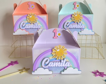 Könnte beinhalten: Drei pastellfarbene Geschenkboxen mit einem Regenbogen-, Sonnen- und Wolkenmotiv. Jede Box trägt den Namen "Camila" und die Zahl "1".