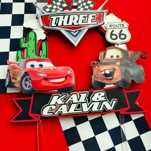 May include: Red and brown Cars themed cake toppers with a checkered flag background. The toppers feature Lightning McQueen and Mater from the Disney Pixar movie Cars. The toppers also include a sign that says "Three" and "Route 66". The toppers are on sticks and are ready to be placed on a cake.