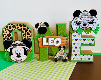 Könnte beinhalten: Ein Satz aus Pappbuchstaben, die "ONE" buchstabieren und mit einem Dschungel-Thema verziert sind. Die Buchstaben sind braun mit grünen und braunen Akzenten. Der Buchstabe "O" hat einen Mickey-Maus-Kopf, der einen Safarihut trägt. Der Buchstabe "N" hat ein Schild mit dem Namen "LEO" darauf. Der Buchstabe "E" hat einen Mickey-Maus-Kopf, der ein Fernglas trägt.