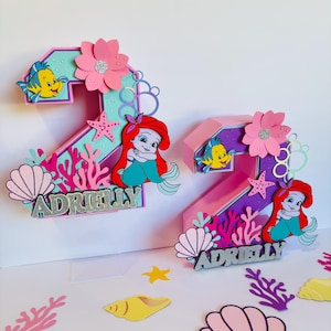 May include: Two pink and purple 3D number two cake toppers with a mermaid and Flounder theme. The toppers are decorated with pink flowers, seashells, coral, and stars. The name "Adrielly" is written on each topper.