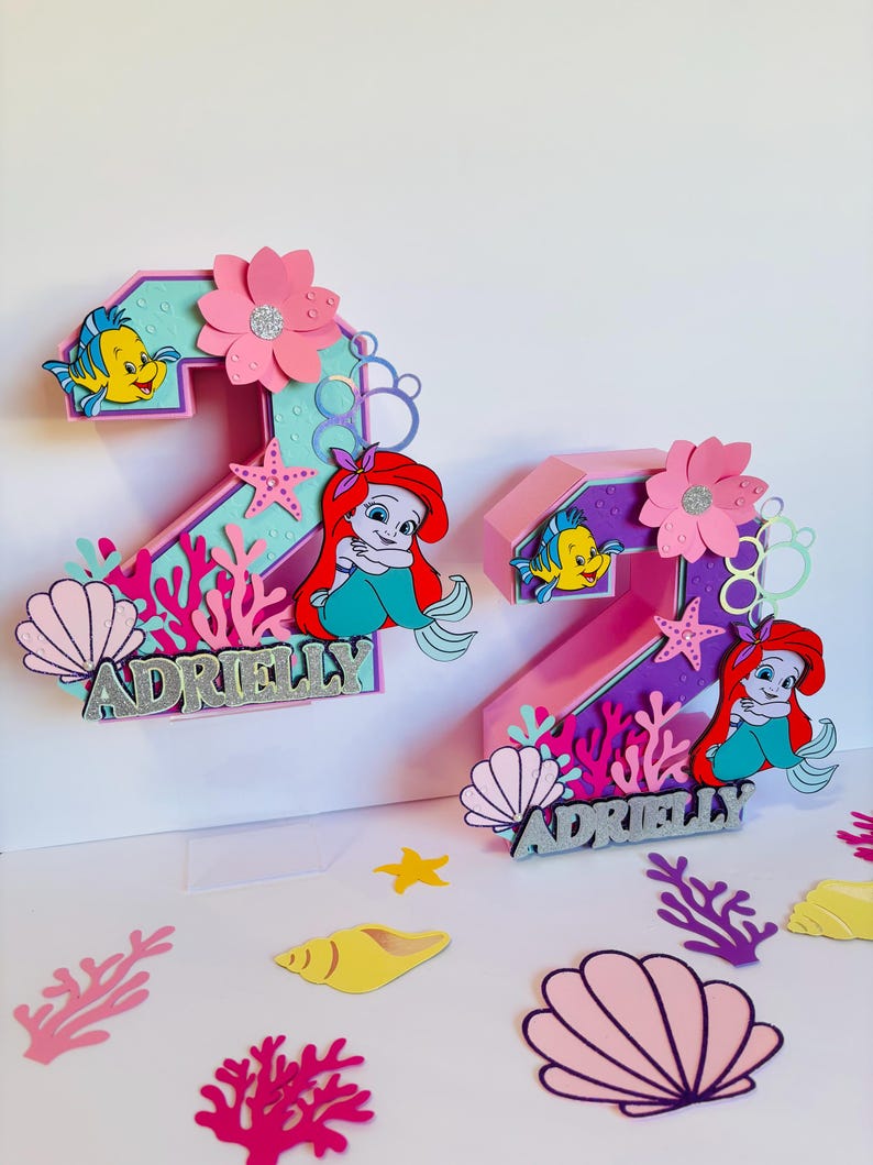 May include: Two pink and purple 3D number two cake toppers with a mermaid and Flounder theme. The toppers are decorated with pink flowers, seashells, coral, and stars. The name "Adrielly" is written on each topper.