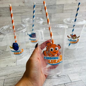 May include: Four clear plastic cups with orange and white striped straws. Each cup has a Finding Nemo themed sticker with a character from the movie. The stickers say "Finding Luka 3".
