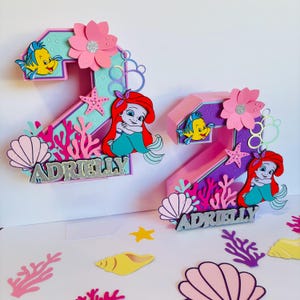 May include: Two pink and purple 3D number two cake toppers with a mermaid theme. Each topper features a mermaid, a starfish, coral, seashells, and the name "Adrielly".