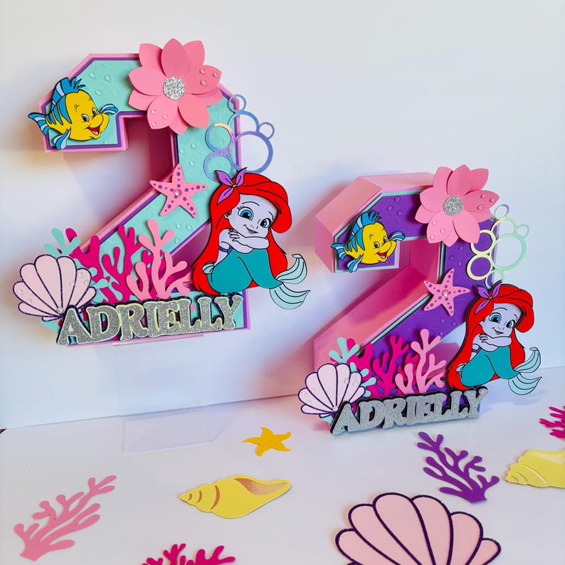May include: Two pink and purple 3D number two cake toppers with a mermaid theme. Each topper features a mermaid, a starfish, coral, seashells, and the name "Adrielly".