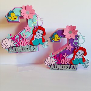 May include: Two purple and pink numbers, 2 and 7, decorated with a mermaid, seashells, and starfish. The numbers are topped with the name "Adrielly" in silver glitter.
