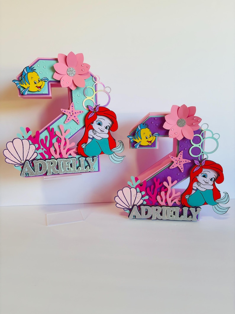 May include: Two purple and pink numbers, 2 and 7, decorated with a mermaid, seashells, and starfish. The numbers are topped with the name "Adrielly" in silver glitter.
