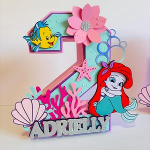 May include: A pink and purple number two cake topper featuring a mermaid, a yellow fish, a pink flower, and a seashell. The topper also includes the name "Adrielly" in silver glitter letters.