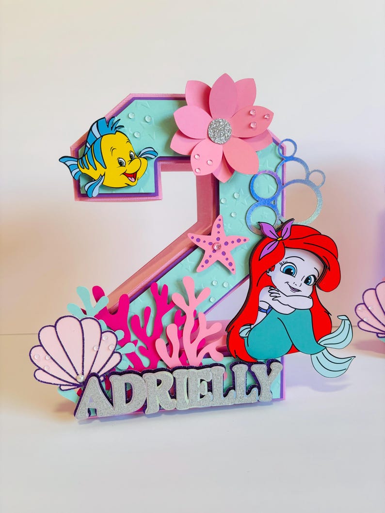 May include: A pink and purple number two cake topper featuring a mermaid, a yellow fish, a pink flower, and a seashell. The topper also includes the name "Adrielly" in silver glitter letters.