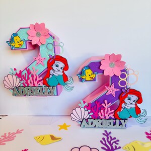 May include: Two pink and purple 3D number two cake toppers decorated with a mermaid, a starfish, seashells, coral, and the name "Adrielly".