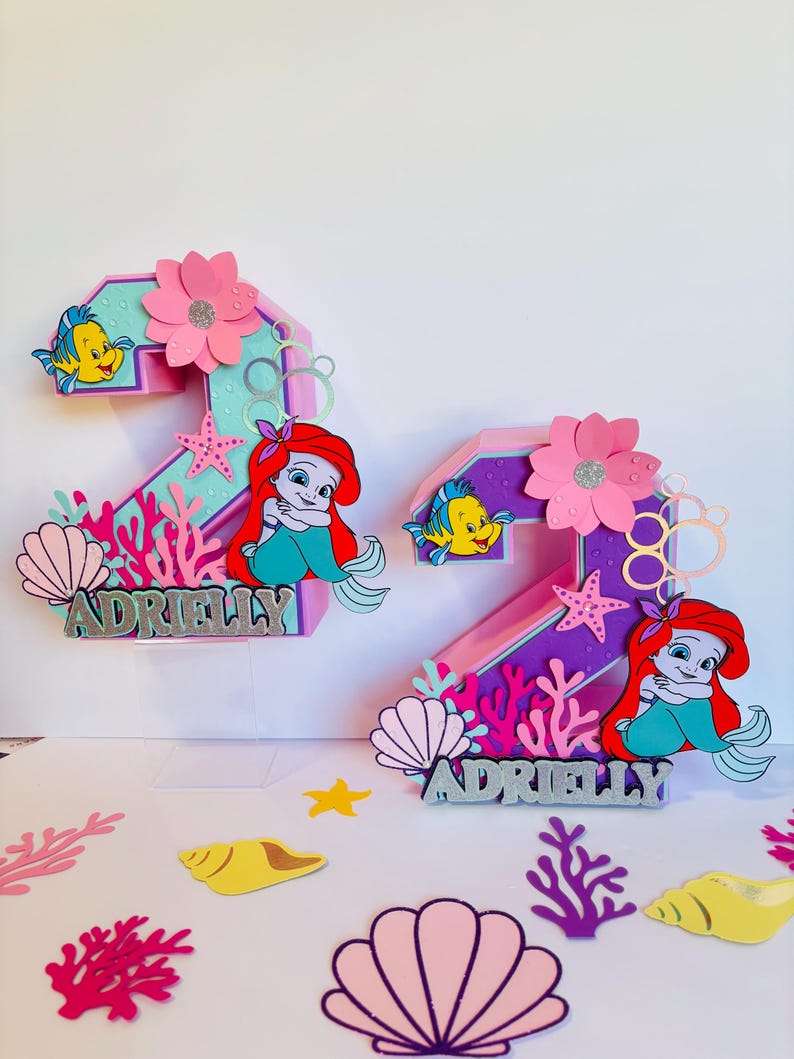 May include: Two pink and purple 3D number two cake toppers decorated with a mermaid, a starfish, seashells, coral, and the name "Adrielly".