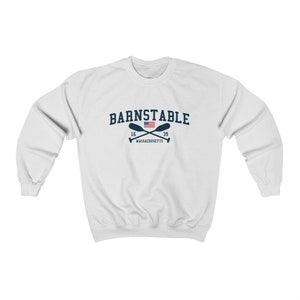 以下が含まれることがあります： 白いスウェットシャツに、ネイビーブルーのグラフィックで「BARNSTABLE」の文字が書かれており、その下に小さなアメリカ国旗と交差したオールが描かれています。「16」と「39」の文字は旗の両側にあり、「MASSACHUSETTS」の文字はオールの下にあります。