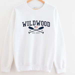 May include: White sweatshirt with a navy blue graphic print of the text "WILDWOOD" with a crossed paddle design and a small American flag. The text "19 12 NEW JERSEY" is printed below the graphic.