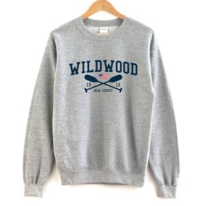 May include: A gray sweatshirt with the text "WILDWOOD" in navy blue lettering. The text is surrounded by two crossed oars with an American flag in the center. The text "19" and "12" are on either side of the oars. The text "NEW JERSEY" is below the oars.